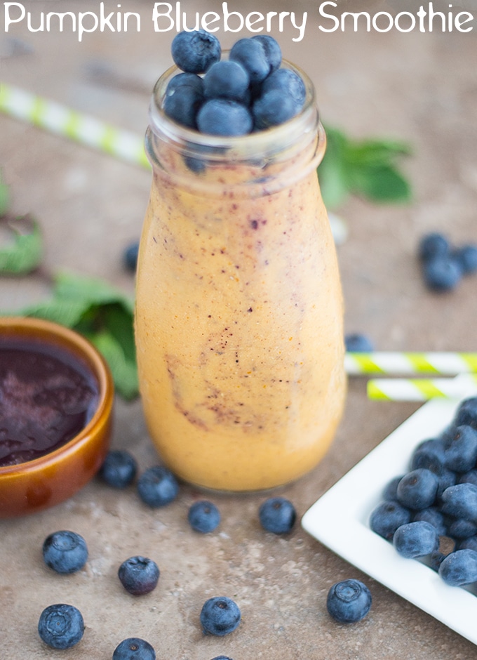 Front View of a Glass Bottle Filled with Pumpkin Smoothie. Its surrounded by Blueberries, Green Stripped Straws, Mint Leaves and a Brown Ramekin with Blueberry Syrup