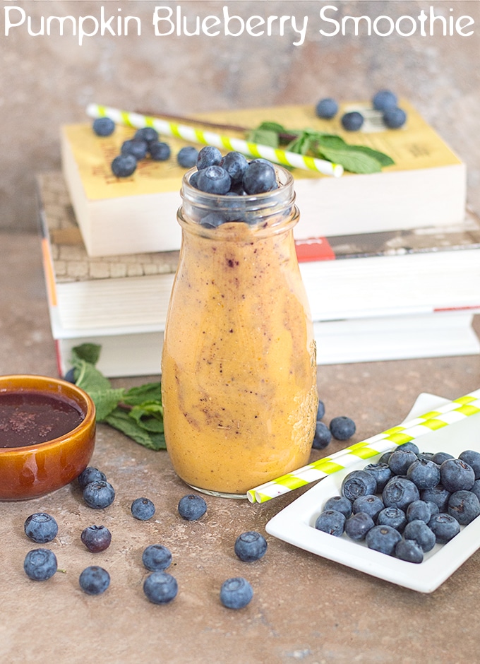 Front View of a Glass Bottle Filled with Pumpkin Smoothie. Its surrounded by Blueberries, Green Stripped Straws, Mint Leaves and a Brown Ramekin with Blueberry Syrup. There is a Stack of Books in the Background
