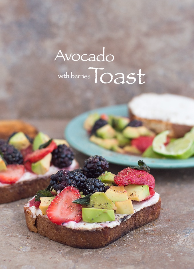 Front View of A Toasted Slice of Bread with Vegan Cream Cheese Spread and Topped with Avocado, Fresh Strawberries and Blackberries. A Similar Toast is in the Background on the Left and a Green Plate with the Fruit Mixture is in the Right Background