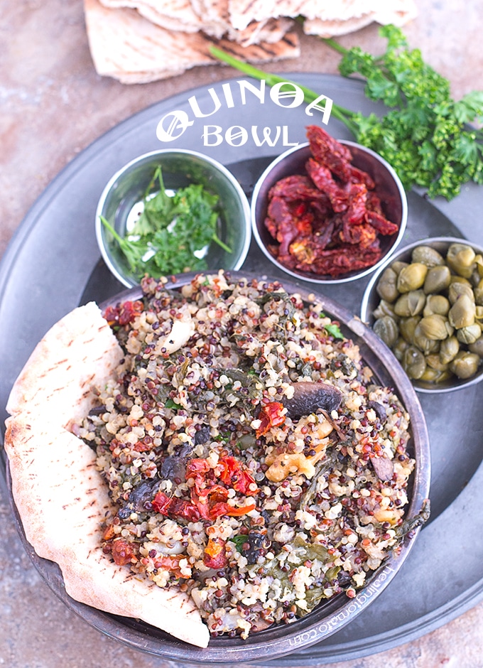 Overhead view of Grey Plate Loaded with 4 Bowls. Main Bowl is Filled with Cooked Quinoa and a 1/2 of a Pita Bread on the Side. 3 Smaller Steel Bowls have Parsley, Sundried Tomatoes and Capers