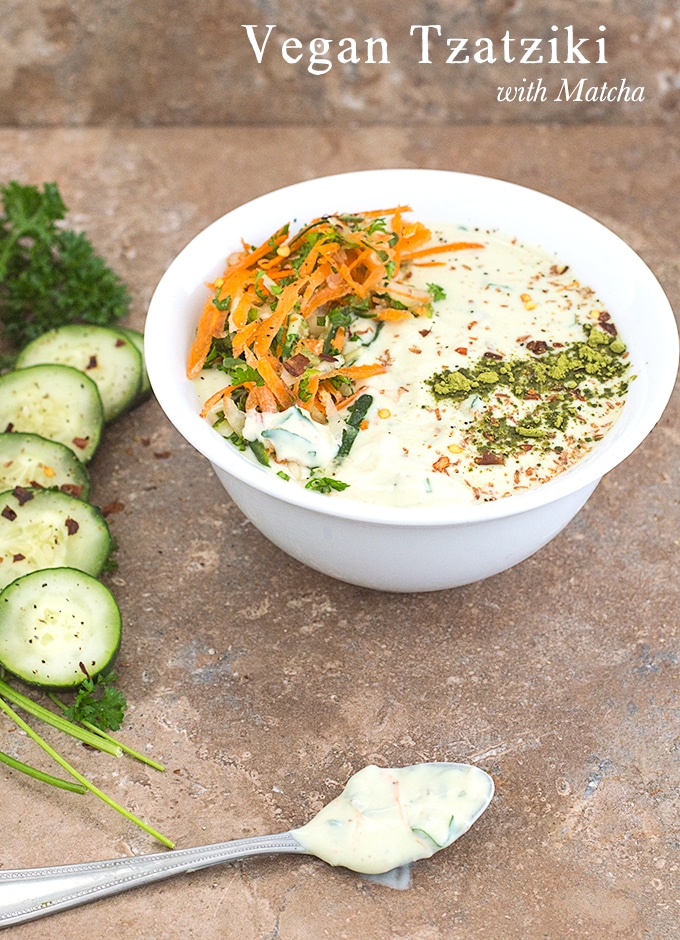 Front view of a white bowl filled with Vegan Tzatziki with carrot saalad on the left and matcha powder line on the right. To the left of the bowl, Parsley, cucumber and lime rounds are placed in an arc