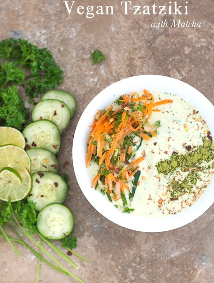 Closeup view of a white bowl filled with Tzatziki with carrot saalad on the left and matcha powder line on the right. To the left of the bowl, Parsley, cucumber and lime rounds are placed in an arc
