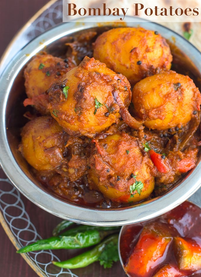 Overhead view of a steel bowl filled with potatoes cooked in Indian Spices. In the Background, curry, naan and papad are visible - Bombay Potatoes recipe