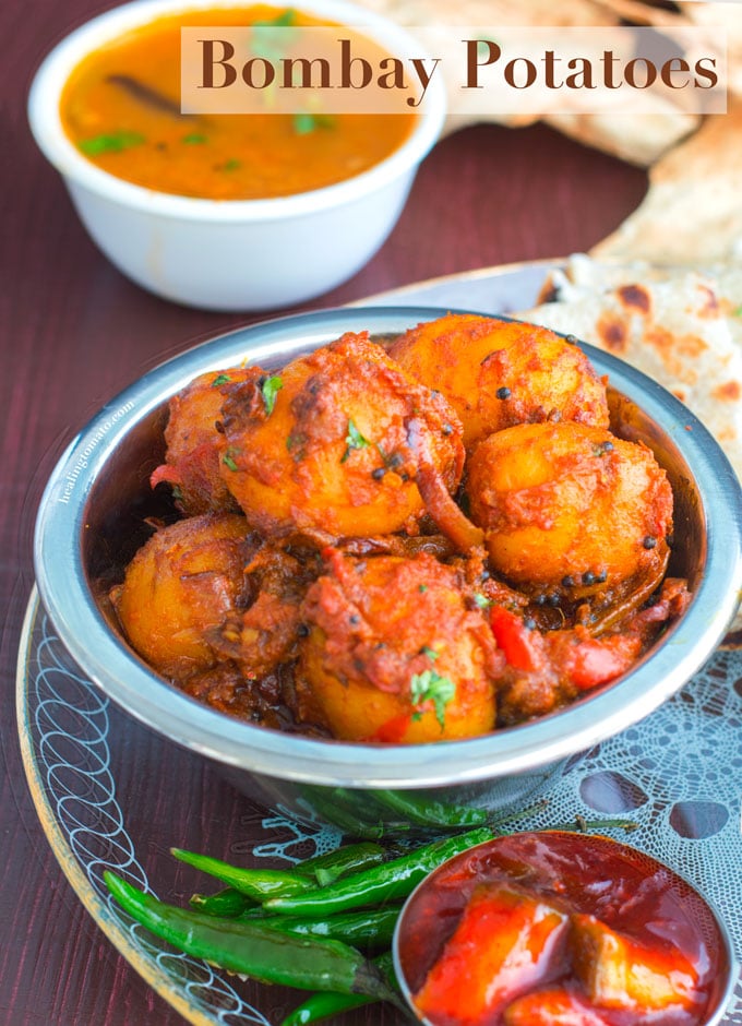 Front view of a steel bowl filled with potatoes cooked in Indian Spices. In the Background, curry, naan and papad are visible - Bombay Potatoes recipe