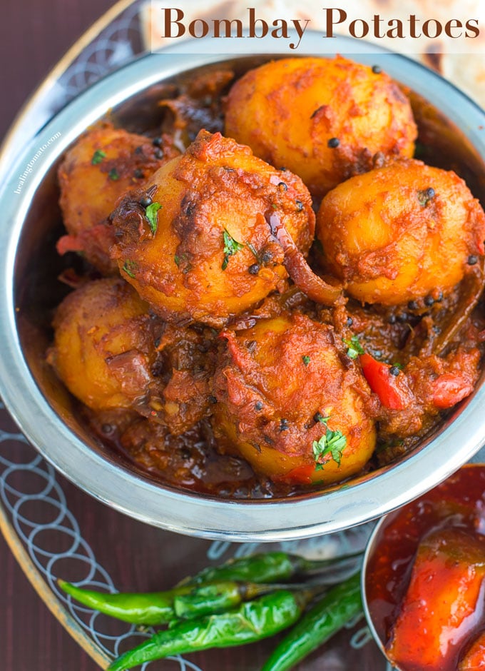 Overhead view of a steel bowl filled with potatoes cooked in Indian Spices. In the Background, curry, naan and papad are visible - Bombay Potatoes recipe
