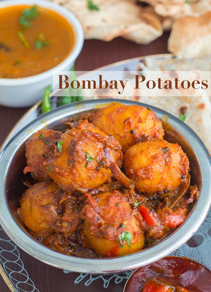 Overhead view of a steel bowl filled with potatoes cooked in Indian Spices. In the Background, curry, naan and papad are visible - Bombay Potatoes