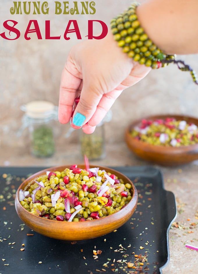Front view of the author's hand sprinkling pomegranate over a brown bowl filled with onions, mung beans and pomegranate