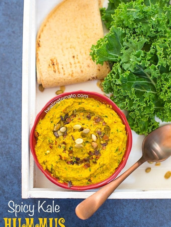 Overhead View of a White Tray with a red bowl in it. Bowl contains Spicy Kale Hummus and is surrounded by Kale leaves, spoon and pita bread