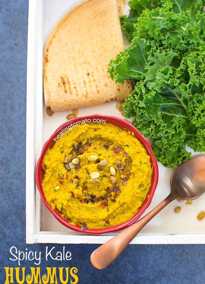 Overhead View of a White Tray with a red bowl in it. Bowl contains Spicy Kale Hummus and is surrounded by Kale leaves, spoon and pita bread