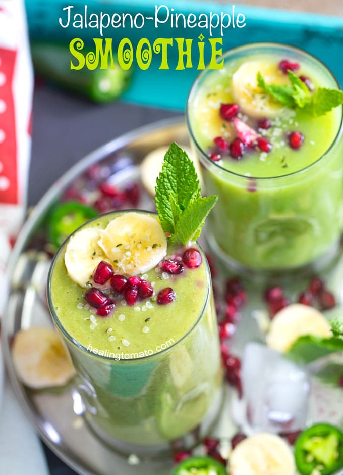 Overhead view of 2 small glasses filled with pineapple smoothie and garnished with banana, mint and pomegranate. They are placed on a stainless steel plate with ice cubes, pomegranate seeds, banana rounds and mint leaves