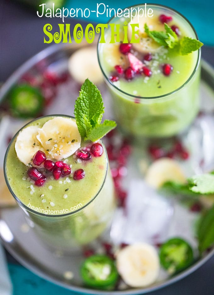 Overhead view of 2 small glasses filled with pineapple smoothie and garnished with banana, mint and pomegranate. They are placed on a stainless steel plate with ice cubes, pomegranate seeds, banana rounds and mint leaves
