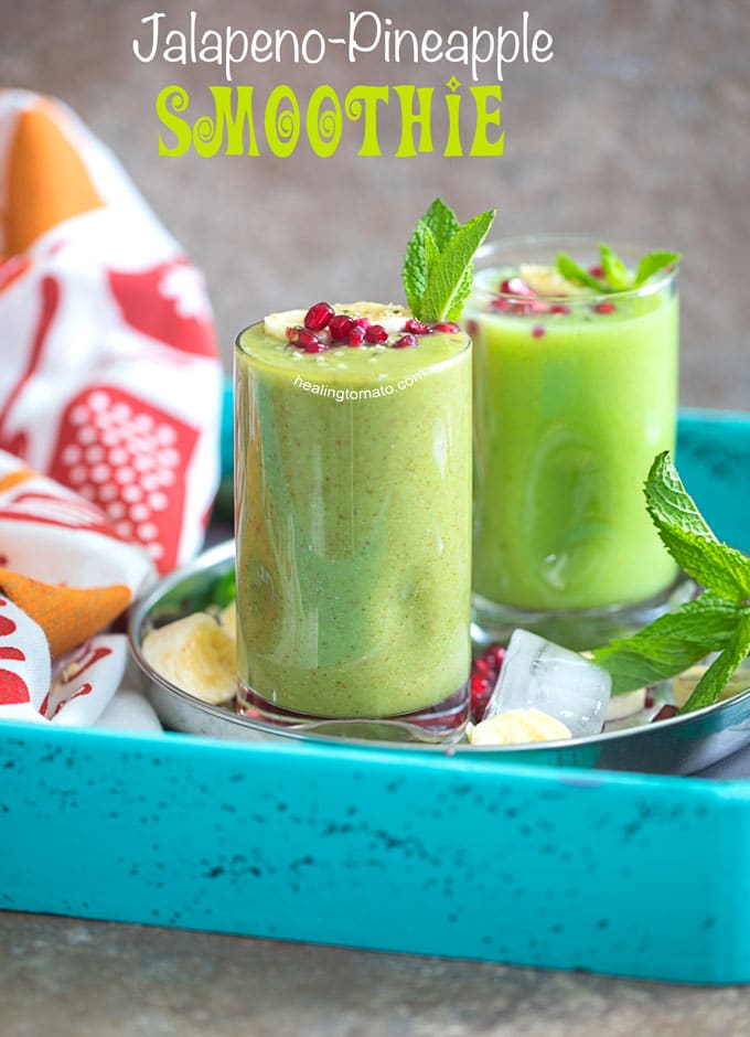 Front view of 2 small glasses filled with pineapple smoothie and garnished with banana, mint and pomegranate. They are placed on a stainless steel plate with ice cubes, pomegranate seeds, banana rounds and mint leaves