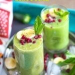 Angle view of 2 small glasses filled with pineapple smoothie and garnished with banana, mint and pomegranate. They are placed on a stainless steel plate with ice cubes, pomegranate seeds, banana rounds and mint leaves