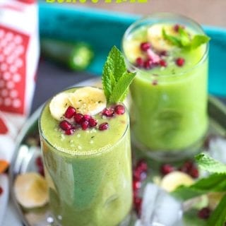 Angle view of 2 small glasses filled with pineapple smoothie and garnished with banana, mint and pomegranate. They are placed on a stainless steel plate with ice cubes, pomegranate seeds, banana rounds and mint leaves
