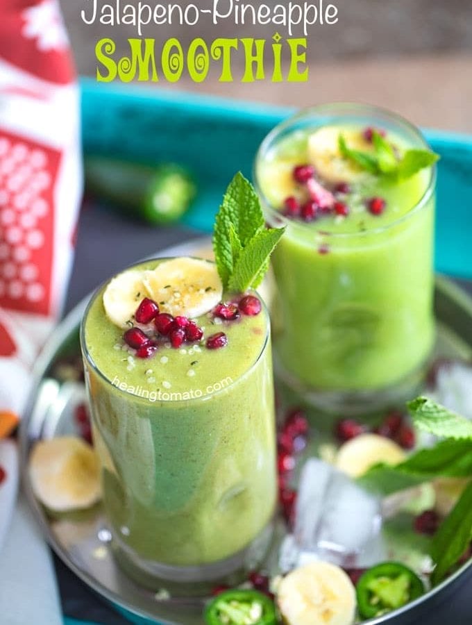 Angle view of 2 small glasses filled with pineapple smoothie and garnished with banana, mint and pomegranate. They are placed on a stainless steel plate with ice cubes, pomegranate seeds, banana rounds and mint leaves