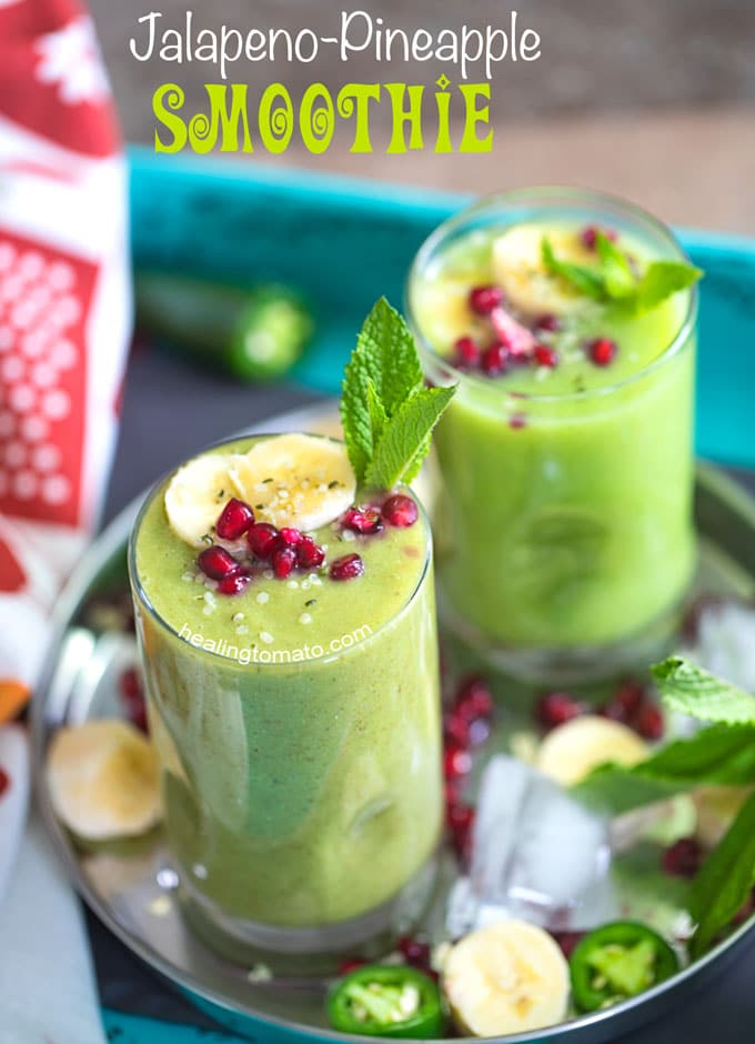 Angle view of 2 small glasses filled with pineapple smoothie and garnished with banana, mint and pomegranate. They are placed on a stainless steel plate with ice cubes, pomegranate seeds, banana rounds and mint leaves