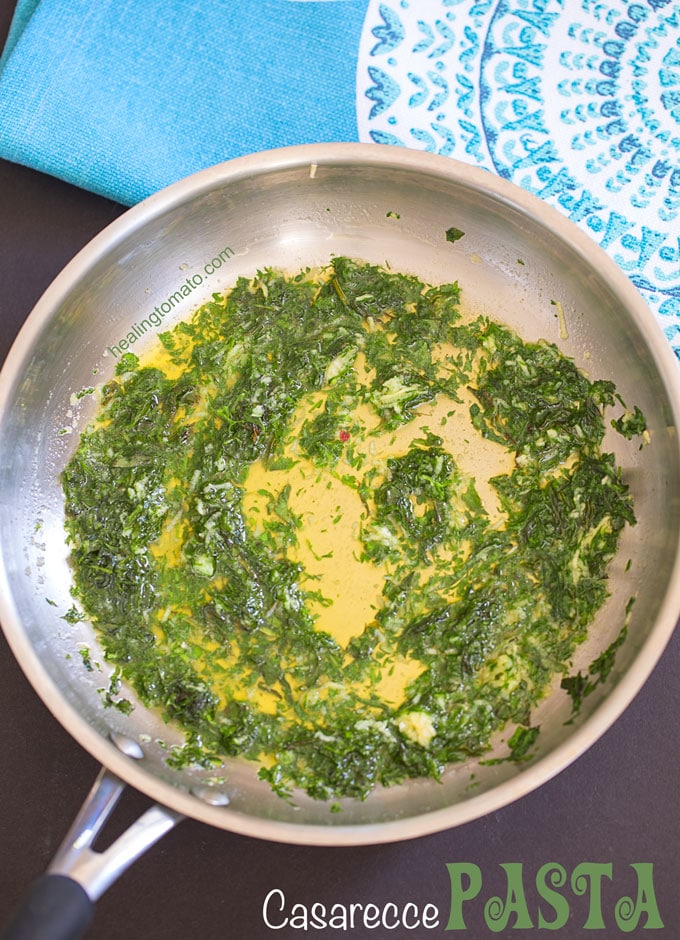 Overhead view of a heavy bottom stainless steel pan filled with a rich vegan butter sauce and chopped fresh herbs. A blue place mat in by its side - casarecce pasta