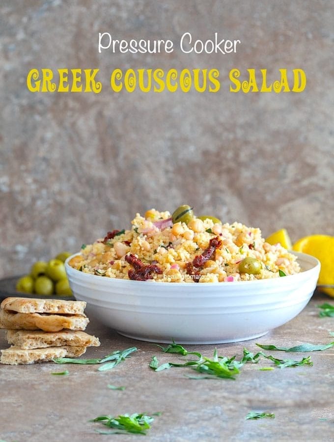 Front view of a white bowl filled with greek couscous sald. Pita bread on the side, olives and lemon rinds in the back