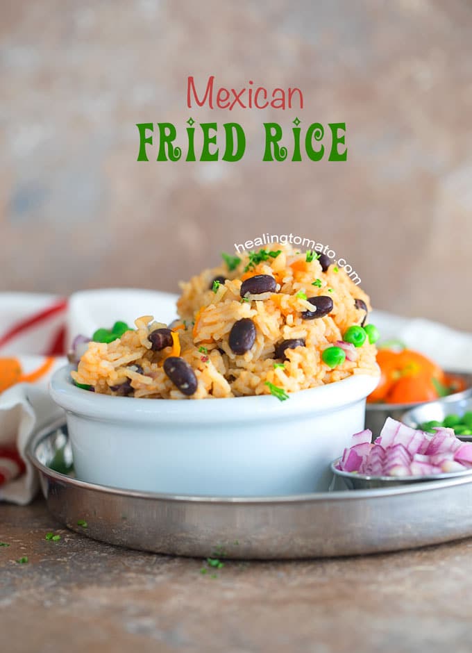 Front View of Mexican Fried Rice in a white bowl with a towel in the background and red onion, peas and tomatoes next to it