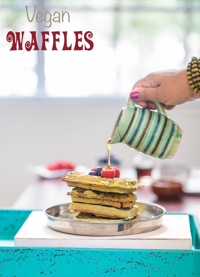 Front view of a stack of 4 kale vegan waffles topped with blueberries and raspberries. A drizzle of maple syrup is being dropped from the top by the author