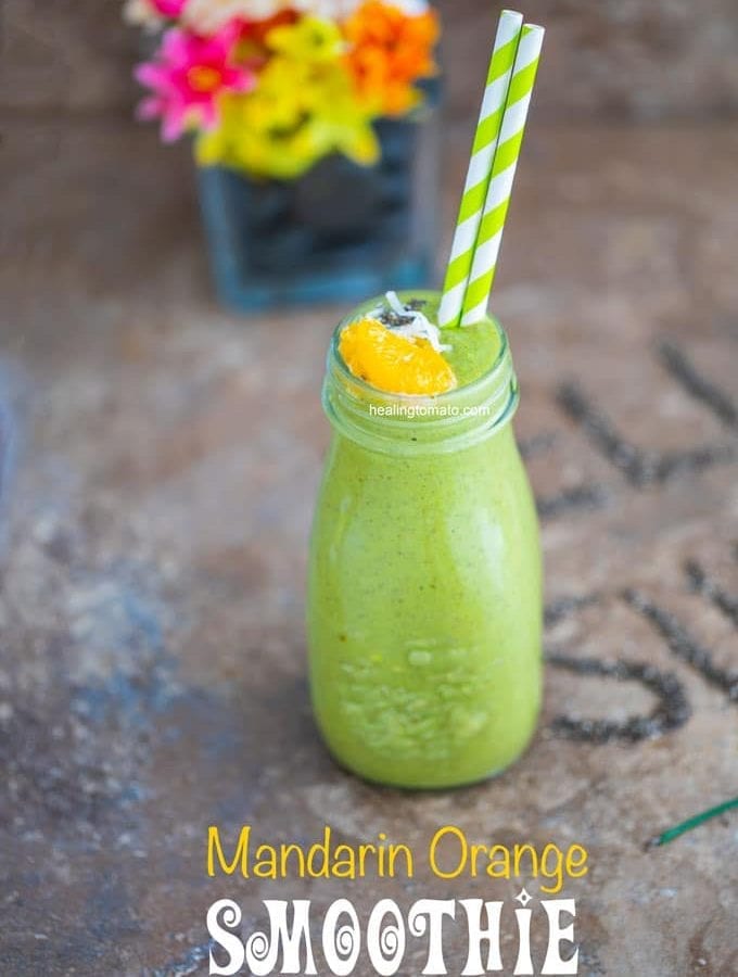 Front view of a glass milk bottle filled with the green smoothie and has 2 plastic straws. Garnished with mandarin oranges and chia seeds. A glass flower vase in the background