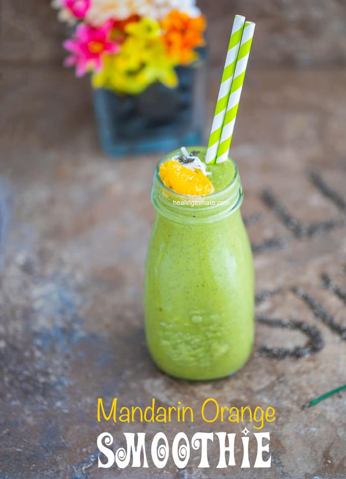 Front view of a glass milk bottle filled with the green smoothie and has 2 plastic straws. Garnished with mandarin oranges and chia seeds. A glass flower vase in the background