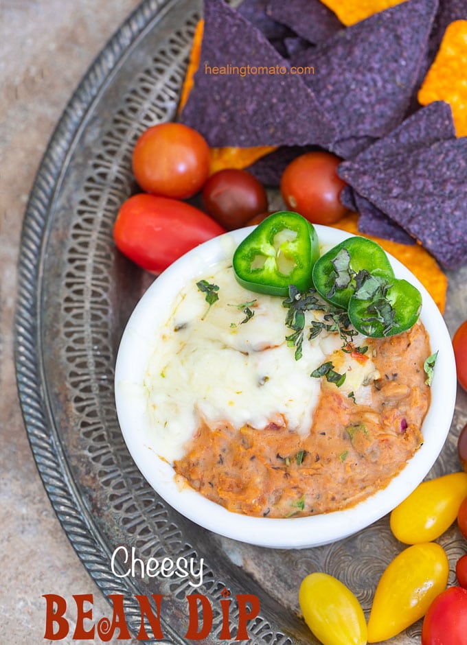 Overhead View of a white bowl filled with mexican bean dip and surrounded by chips and cherry tomatoes. They are all placed on a grey tray