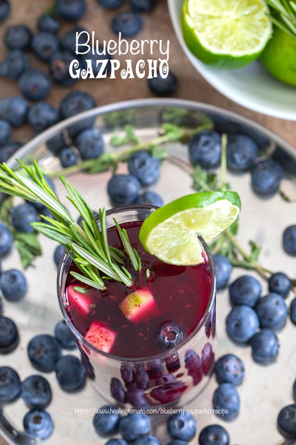 Overhead view of a glass filled with Gazpacho soup and garnished with Lime and Rosmary Stem. Glass surrounded by blueberries, lime and flowers