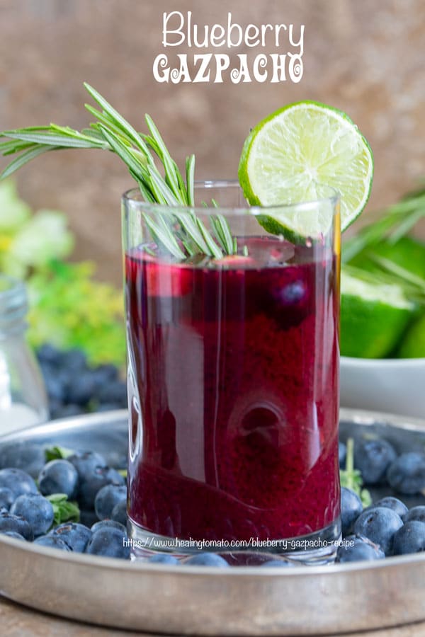 Front view of a glass filled with Gazpacho soup and garnished with Lime and Rosmary Stem. Glass surrounded by blueberries, lime and flowers