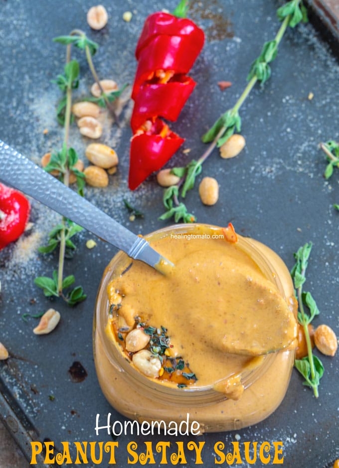 Overhead view of a spoon filled with the peanut satay sauce resting over the top of the mason jar. The jar is on a baking tray filled with red chili pepper, oregano stems, peanuts and onion powder