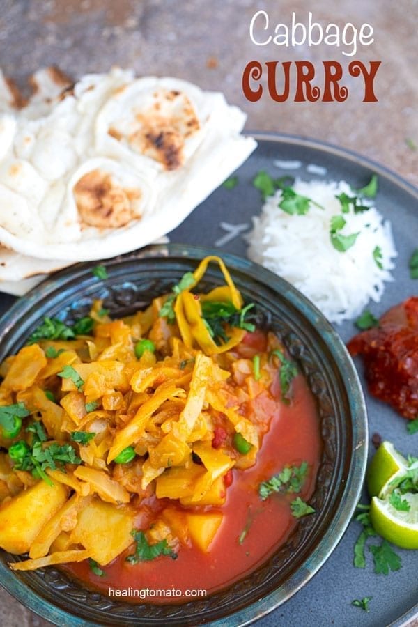 Overhead view of a metallic grey bowl filled with cabbage curry in a red sauce. Bowl surrounded by naan, rice, pickels and lime - Vegan Cabbage Curry