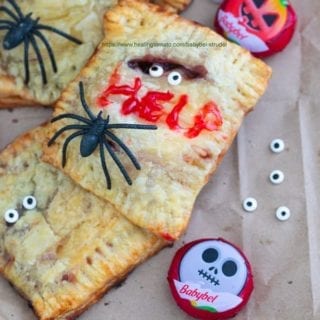 Closeup view of a Babybel strudel surrounded by Babybel cheese, candy eyes and spiders. One of the strudels has the word “HELP” written with icing - Babybel Strudel