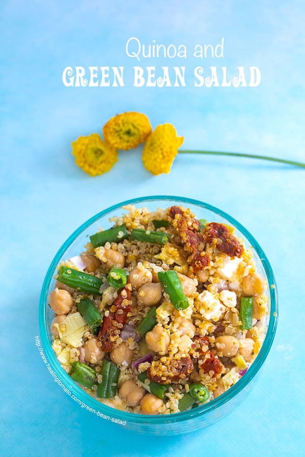 Closeup view of a bowl on a blue background. Bowl is filled with quinoa, green beans and chick peas. Garnished with cilantro and orange slices. Small yellow carnations on the side - Green Bean Salad