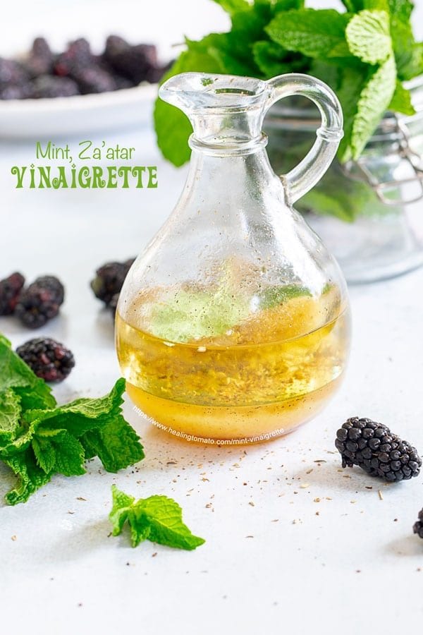 Front view of a small glass cruet filled with mint vinaigrette. Background has mint and blackberry