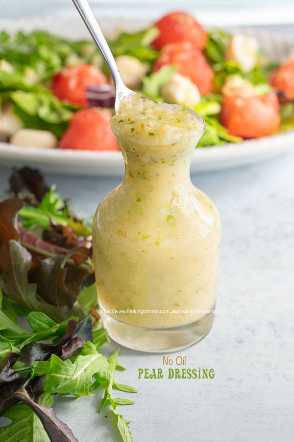 Front view of a small glass bottle filled with pear dressing with arugula salad in the background