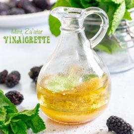 Front view of a small glass cruet filled with mint vinaigrette. Background has mint and blackberry