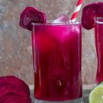 Front view of a small glass filled with beet lemonade. Ice cubes inside, a red paper straw to the side and a slice of beet lemonade for garnish