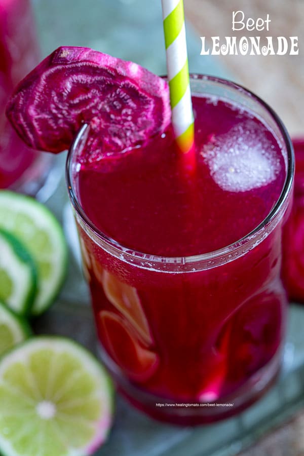 Closeup view of 1 small glasses filled with beet lemonade. Ice cubes inside, a green paper straw to the side and a slice of beet lemonade for garnish