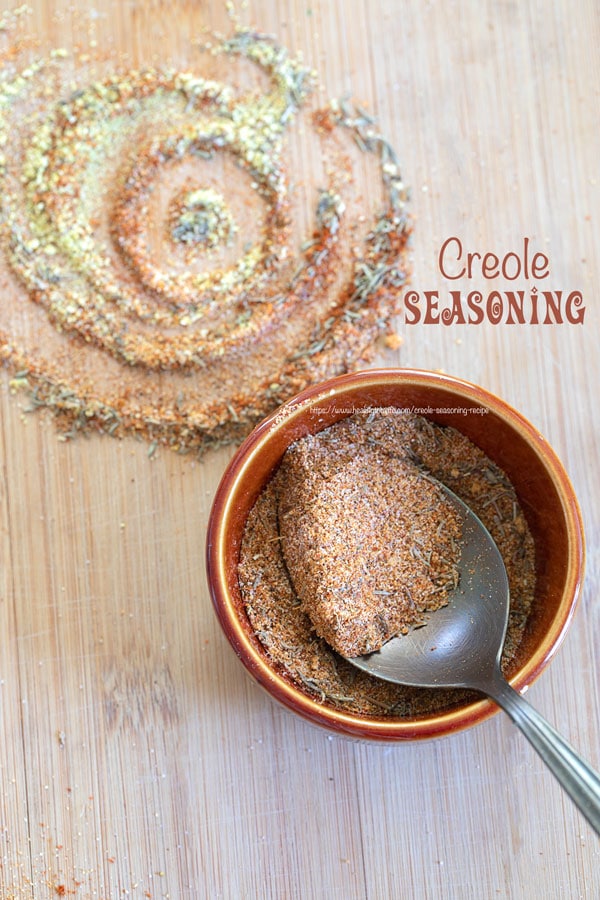 Top view of a small brown bowl filled with creole seasoning with a spoon in the bowl. Next to the bowl, there is a spiral shape of some of the powdered ingredients