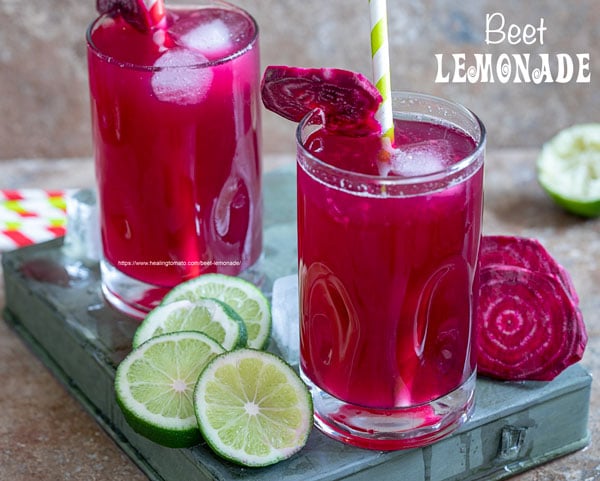 Front view of two small glasses filled with beet lemonade. Ice cubes inside, red, green paper straw to the side and a slice of beet lemonade for garnish