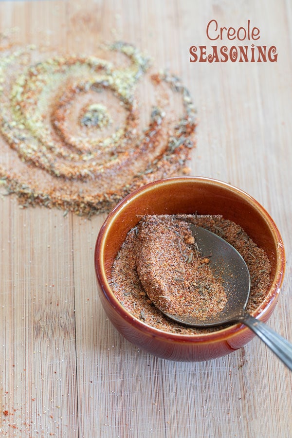 Front view of a small brown bowl filled with creole seasoning with a spoon in the bowl. Next to the bowl, there is a spiral shape of some of the powdered ingredients