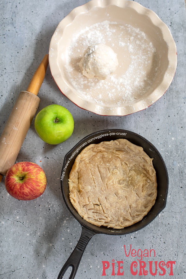 Top view of a pie crust in a cast iron pan. A rolling pin, apples, and pie pan on the side