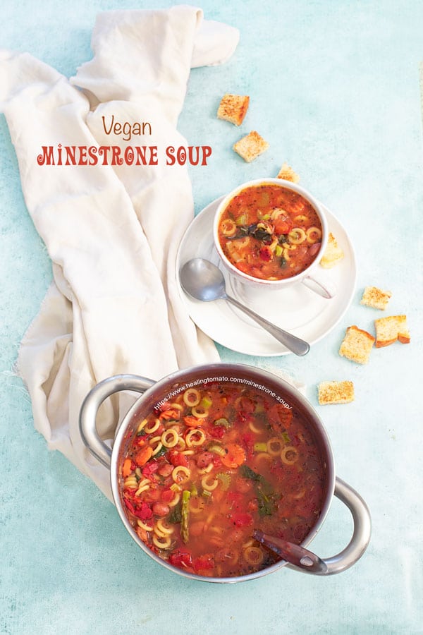 Top view of a stainless steel pan filled with minestrone soup. On the side, a cup filled with minestrone soup is on a plate and croutons strewn about