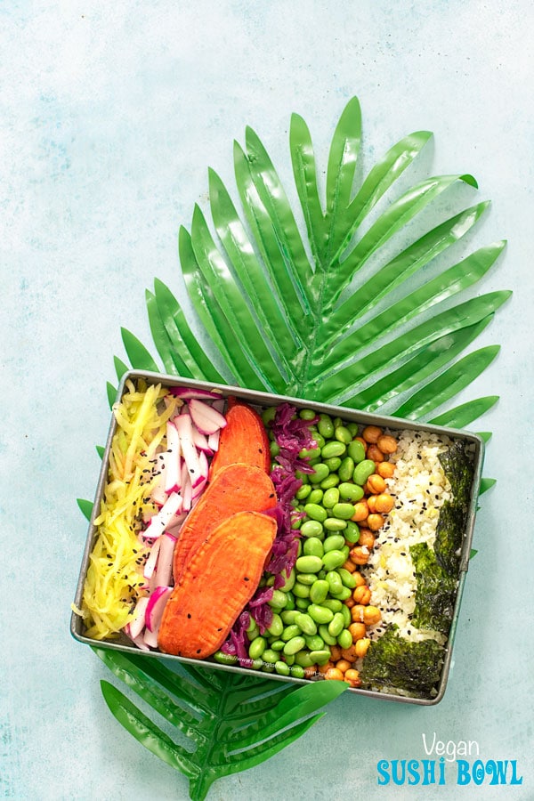 Top view of a rectangle metal tray on a long palm leaf. Metal tray has the following ingredients arranged in a row: shredded mango, radishes, potatoasts, red cabbage kraut, edamame, chickpea snack, riced cauliflower and seaweed squares