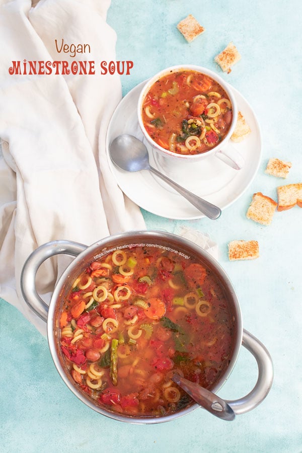 Top view of a stainless steel pan filled with minestrone soup. On the side, a cup filled with minestrone soup is on a plate and croutons strewn about