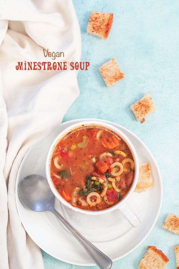 A top view of a cup filled with minestrone soup is on a plate and croutons strewn about