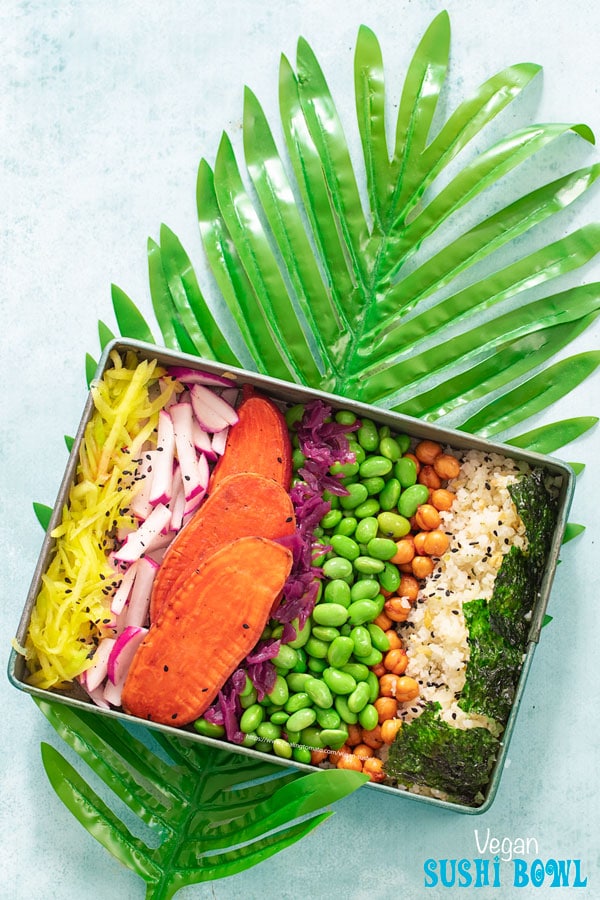 Top view of a rectangle metal tray on a long palm leaf. Metal tray has the following ingredients arranged in a row: shredded mango, radishes, potatoasts, red cabbage kraut, edamame, chickpea snack, riced cauliflower and seaweed squares