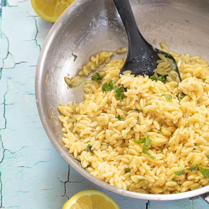 Top view of a stainless steel pan filled with cooked lemon orzo pasta and 2 lemon halves on the side of the pan