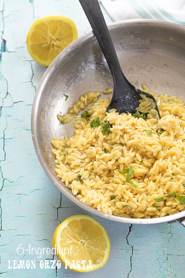 Top view of a stainless steel pan filled with cooked lemon orzo pasta and 2 lemon halves on the side of the pan