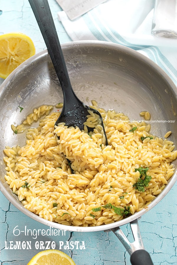 Top view of a stainless steel pan filled with cooked lemon orzo pasta with a black spoon in the pan, lemon halves on the outside, a cloth and a glass bottle with salt